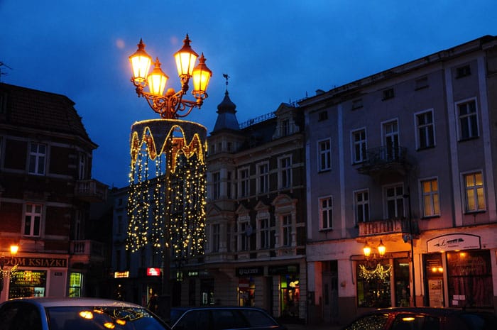 Ozdobna trójramienna latarnia na rynku, spod kloszy spływa złota kurtyna świetlna; w tle kamienice o zmierzchu