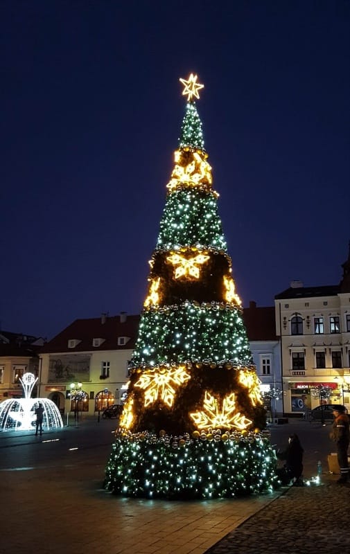 Nocne zdjęcie realizacji: wysoka choinka stożkowa z gwiazdą i złotymi śnieżynkami na rynku, obok podświetlona fontanna